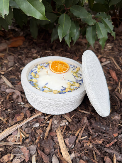 Betonkerze mit Deckel "Grey Secret" (12,5cm) aus hellgrauem Beton. Wiederverwendbare Dose mit 2 Baumwolldochten und Sojawachs. Dekoriert mit einer getrockneten Orangenscheibe und blauen Blütenblättern.