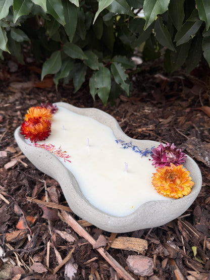 Design-Betonkerze "Grey Wave" in organischer Wellenform, 29,5cm lang. Hellgraue Betonschale gefüllt mit veganem Sojawachs und drei Baumwolldochten. Dekoriert als "Flower Edition" mit bunten Strohblumen in Orange und Rot sowie blauen Blütenblättern. Handgegossen im Schwarzwald.