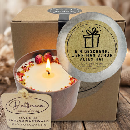 Brennende Vanille-Duftkerze mit Rosenknospen und Blütenblättern in Vintage-Dose vor Geschenkbox mit Deckelspruch